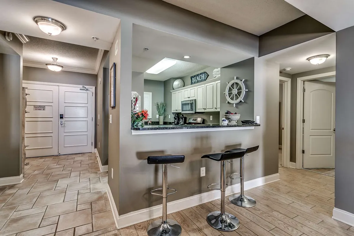A welcoming view of the kitchen area featuring bar stools at the counter, modern appliances visible in the background, and beach-themed decor throughout. The image captures a sense of sociability and culinary capability, with ample space for prepping and entertaining.