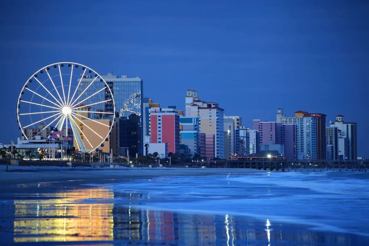 The image features a colorful Ferris wheel, vibrant buildings, and ocean waves, all seen from the beach. It highlights a playful, family-friendly atmosphere, with opportunities for relaxation and fun by the water's edge. The sunset lighting and beachfront setting evoke the joy and beauty of the coastline.