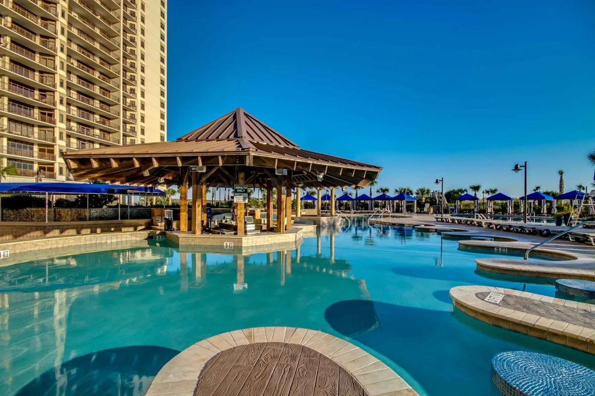 A lively outdoor pool area highlighted by a sparkling pool, comfortable lounge chairs, and bright blue umbrellas. A pavilion is visible, suggesting a space for social gatherings or events. The scene exudes a leisurely mood, with guests sunbathing, socializing, and enjoying drinks by the water.