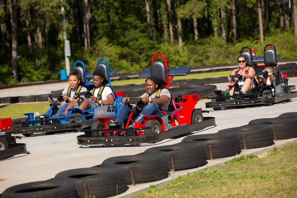 An outdoor go-kart racing track surrounded by greenery, with participants actively racing and spectators enjoying the action. The environment is filled with laughter and friendly competition, ideal for families and groups.