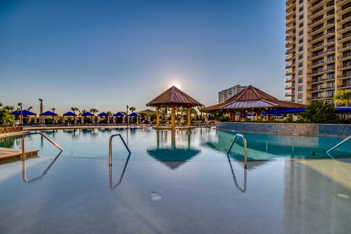 A serene sunset scene at the resort’s swimming pool, with thatched-roof cabanas and warm evening light. Guests are seen relaxing by the pool, socializing, and enjoying the transition from day to evening in a resort setting.