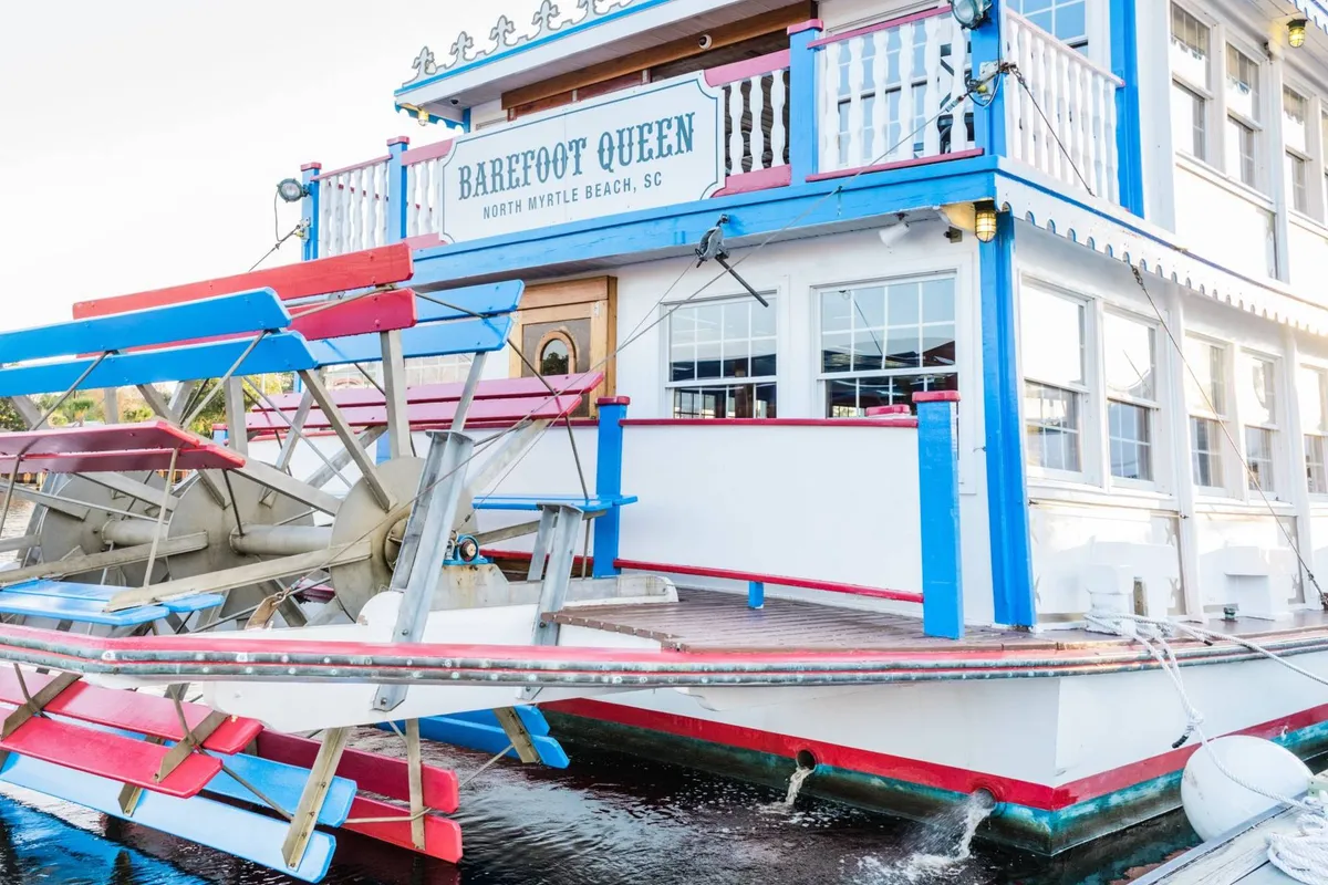 This image depicts the 'Barefoot Queen,' a paddlewheel boat with a colorful exterior, docked by the water. The atmosphere is casual and inviting, with the promise of sunset cruises, live music, and themed dining experiences. The boat suggests opportunities for exploration and adventure along the coast.