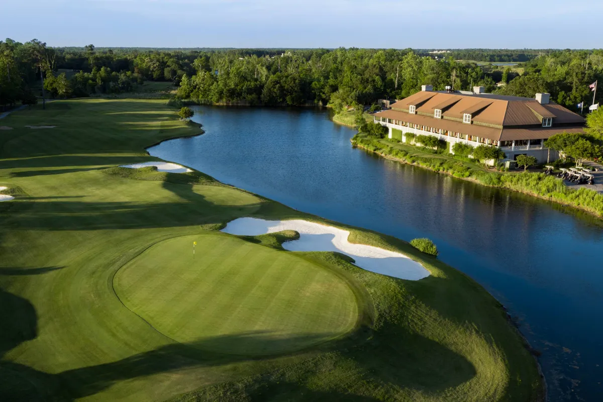 A scenic exterior view of a lush golf course featuring a well-manicured green, a spacious clubhouse, a sparkling lake bordered by sand traps, and ample open sky. The area is ideal for golf enthusiasts and also offers inviting spaces for outdoor dining and social gatherings.