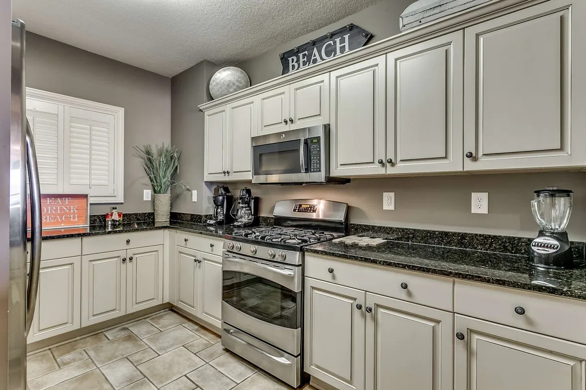 A bright, relaxed kitchen scene showcasing stainless steel appliances, granite countertops, and a prominent blender on the counter. Decorative touches, like a 'BEACH' sign, create a cheerful vibe, while the space clearly supports cooking, baking, and entertaining.