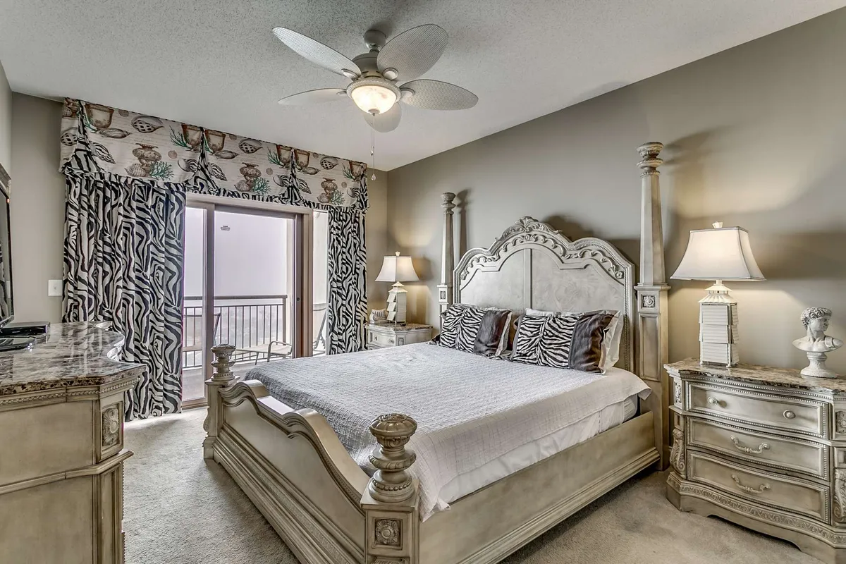 A tranquil bedroom featuring a four-poster bed, zebra-patterned pillows, and sliding glass doors that open directly onto the balcony. Natural light pours in, and the scene hints at both relaxation and easy access to the oceanfront view.